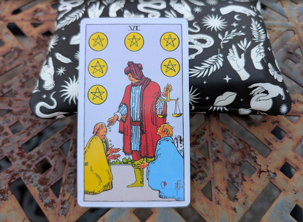 The Six of Pentacles is propped up on a small black and white bag. Both of them are sitting on a rusty patio table.